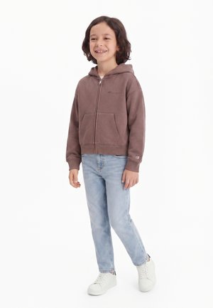 Lachend kind met donker schouderlang haar, een bruine zip-hoodie, lichtblauwe spijkerbroek en witte sneakers, staand tegen een witte achtergrond.