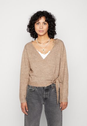Beige indpakket sweater med lange ærmer, skrå halsudskæring og bånddetalje i taljen. Båret over en hvid top og parret med mørkegrå jeans.