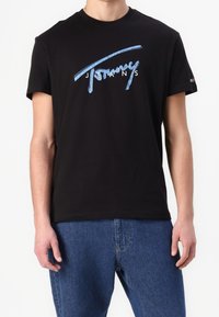 Černé bavlněné tričko s modrým grafickým logem, které říká "Tommy Jeans." Krátké rukávy a kulatý výstřih. Ležérní střih.