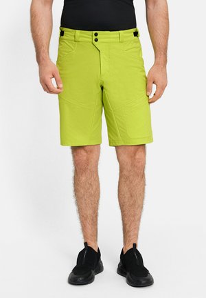 Mann trägt limettengrüne knielange Shorts und schwarze Slip-On-Sneaker, steht vor einem schlichten weißen Hintergrund.