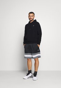 Felpa con cappuccio nera, pantaloni corti sportivi grigi con strisce bianche e scarpe sportive bianco/nero con motivo. Completo sportivo casual.