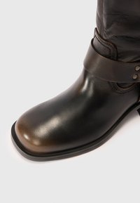 Acercamiento de una bota de cuero marrón con punta redondeada y una correa decorativa alrededor del tobillo sobre un fondo blanco.