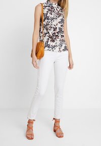Květinová bezrukávová blůza s vysokým límcem, bílé bavlněné džíny a oranžové sandály na řemínky. Hnědá crossbody taška s nastavitelným popruhem.