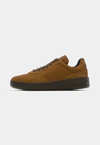 Brown suede sneakers with a textured finish, dark laces, and a chunky sole. Features a logo on the heel and side panel.