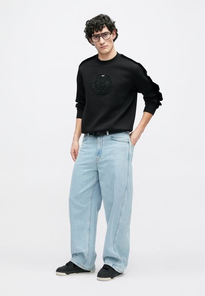 Melns džemperis ar teksturētu apaļu logotipu krūtīs, kombinēts ar gaiši ziliem plaša piegriezuma džinsiem un melnām sporta kurpēm.