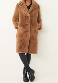 Manteau en fausse fourrure brune avec un col cranté, une fermeture à l'avant et des poches latérales, associé à des leggings noirs en simili cuir et des bottes noires épaisses.