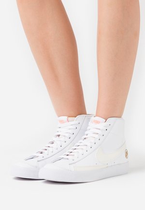 Hvide Nike høje sneakers med snørebånd, båret på fødderne, med lyserød branding og beige swoosh, mod en ensfarvet hvid baggrund.