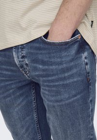 Vaqueros de mezclilla en azul oscuro con una textura ligeramente desgastada, que presentan un diseño tradicional de cinco bolsillos y un pespunte marrón en contraste.