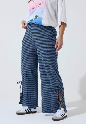 Persona che indossa pantaloni a gamba larga a righe blu e nere con spacchi laterali annodati a fiocco, maglietta bianca con grafica e sneakers bianche Adidas.