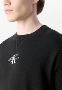 Black knit sweatshirt with a round neckline, featuring textured shoulder accents and an embroidered Calvin Klein logo in white.