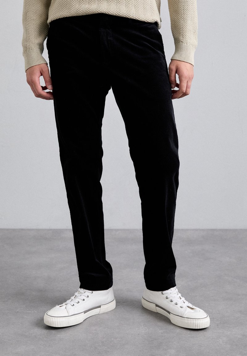 Pantalon en velours côtelé noir avec une coupe droite, associé à des baskets blanches avec un accent argenté, créant une apparence texturée.