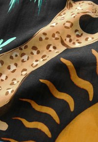 Tissu à motif présentant un léopard bondissant beige à taches brunes sur un fond noir, agrémenté de volutes jaunes évoquant le soleil et de feuillage vert.