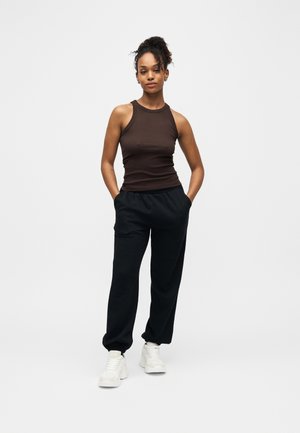 Jeune femme avec un chignon bouclé portant un haut marron sans manches, un pantalon de jogging noir et des baskets blanches, debout les mains dans les poches devant un fond blanc.