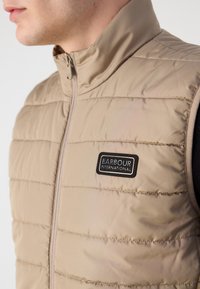 Μπεζ αμάνικο γιλέκο με ψηλό γιακά, κλείσιμο με φερμουάρ και λογότυπο Barbour International. Διαθέτει οριζόντια ραφή για υφή.