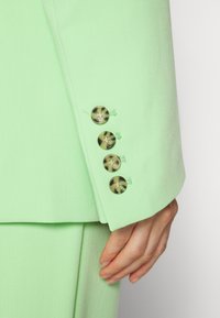 Main reposant sur le côté portant une veste de costume vert clair avec quatre boutons en écaille de tortue marron sur le poignet.