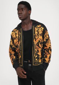 Homme portant un hoodie zippé à motifs noirs et dorés sur un débardeur noir et un jean noir, se tenant contre un fond blanc uni.
