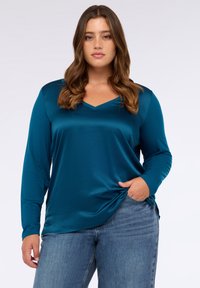 Tyrkysová saténová blůza s dlouhými rukávy, V-výstřihem a bočním rozparkem, v kombinaci s modrými džíny, zdůrazňující hladkou texturu a volný střih.