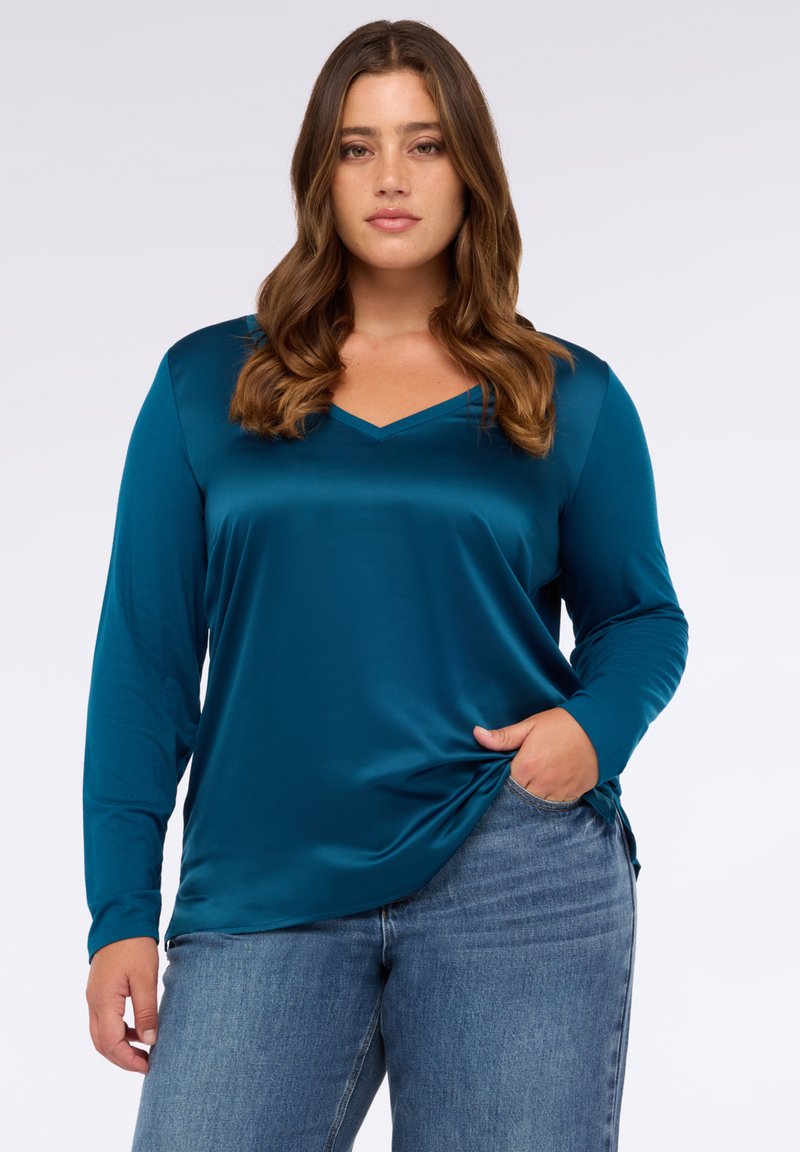 Tyrkysová saténová blůza s dlouhými rukávy, V-výstřihem a bočním rozparkem, v kombinaci s modrými džíny, zdůrazňující hladkou texturu a volný střih.