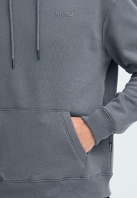 Sweat-shirt gris avec capuche à cordon, poche kangourou et poignets côtelés. Présente un logo "blend" discret sur la poitrine. Texture douce.