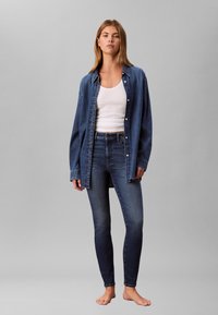 Tamsiai mėlyna džinsinė marškinėliai su sagų užsegimu ant baltos liemenėlės, derinami su aptemptomis tamsiai mėlynomis džinsais. Modelis stovi basas prieš pilką foną.