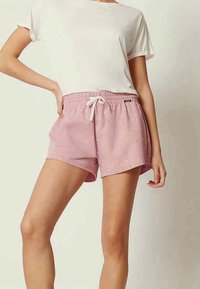 Kvinna som bär rosa dragsko-short med små vita och blå tryck och en vanlig vit t-shirt med kort ärm, står med ena handen på höfterna.