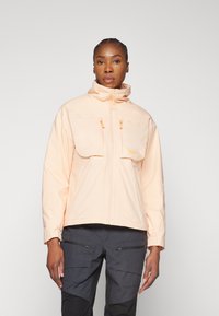 Veste waterproof pêche clair avec un col haut, deux grandes poches avant et des tirettes de zip jaunes contrastantes, assortie à un pantalon cargo noir.