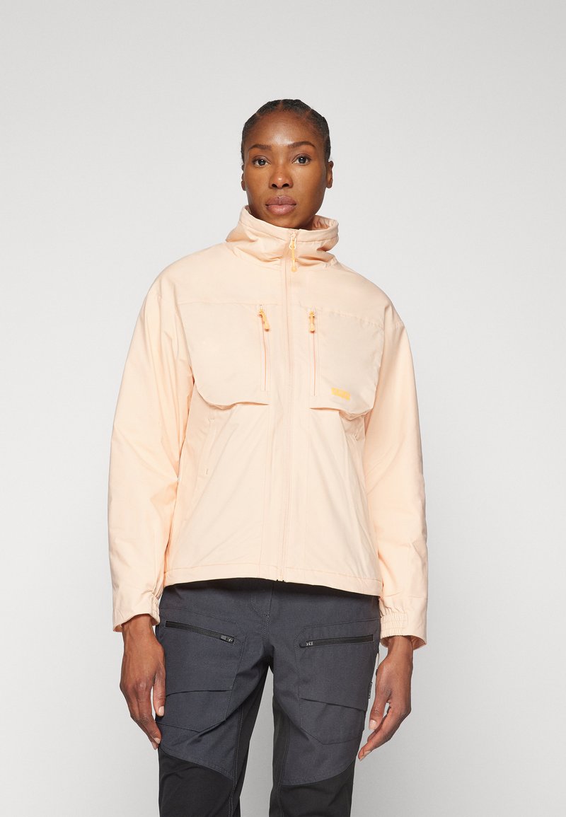 Veste waterproof pêche clair avec un col haut, deux grandes poches avant et des tirettes de zip jaunes contrastantes, assortie à un pantalon cargo noir.