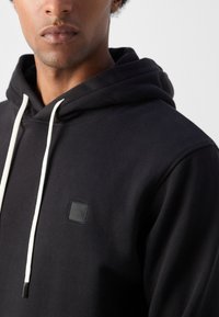 Hoodie noir avec une poche kangourou à l'avant, un cordon blanc, et un patch logo noir carré sur la poitrine. Tissu doux et texturé.