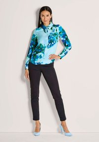 Woman wearing a blue floral button-up blouse, black slim pants, blue high heels, and green earrings, standing with one hand on her hip.