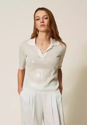 Giovane donna con capelli rossastri che indossa un top in maglia bianco con collo e pantaloni bianchi plissettati a vita alta, in piedi con le mani nelle tasche.