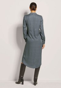 Langarmkleid in Marineblau mit weißen kreisförmigen Mustern, mit geradem Schnitt und hohem Schlitz, kombiniert mit kniehohen schwarzen Lederstiefeln.