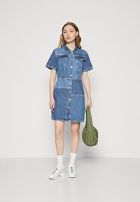 Denim srajčna obleka s kratkimi rokavi, v svetlo in temno modri barvni kombinaciji, sprednjim gumbom in dvema žepoma na prsih. Model drži zeleno torbo.