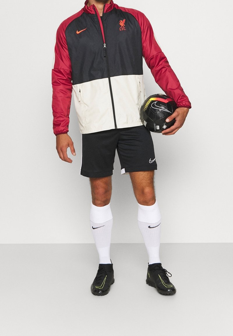 Personne portant une veste Liverpool FC noire, rouge et blanche, un short noir, des chaussettes blanches Nike, des crampons noirs, tenant un ballon de football noir Nike.
