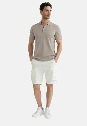 Uomo con polo beige, pantaloncini cargo bianchi e sneakers bianchi, in piedi con le mani nelle tasche, guarda di lato.