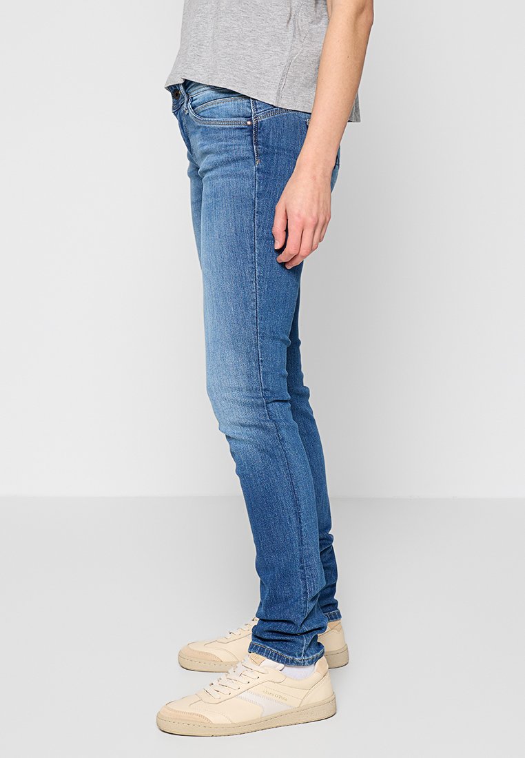 Schmale Jeans aus hellblauem Denim. Verfügt über ein klassisches Fünf-Taschen-Design mit subtilen Verwaschungen, kombiniert mit Sneakers in neutralen Farben.