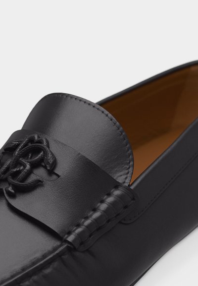 Μαύρο δερμάτινο μοκασίνι με ραμμένα άκρα και διακοσμητικό συνδεδεμένο λογότυπο στο λουράκι, με ταμπά εσωτερική επένδυση.