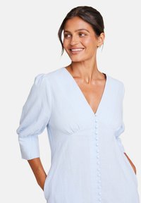 Lys blå linnekjole med dyp V-hals, puffede ermer og knepping foran. Har sidelommer og en løs passform.