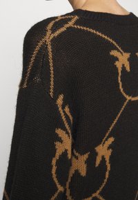 Pull en maille noire avec un motif floral abstrait marron, montré sur l'épaule et le haut du dos d'une personne sur un fond uni.