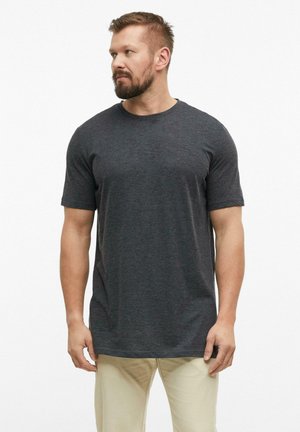 Man met baard die een donkergrijs T-shirt met korte mouwen en beige broek draagt, kijkt naar links tegen een effen witte achtergrond.
