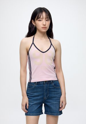 Kvinde iført lyserød haltertop med nummerprint og mørkeblå denimshorts, stående mod en ensartet baggrund.