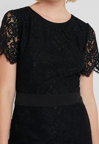 Robe en dentelle noire avec des manches courtes ornées de dentelle, un col rond et une taille lisse. Le tissu présente une texture florale.