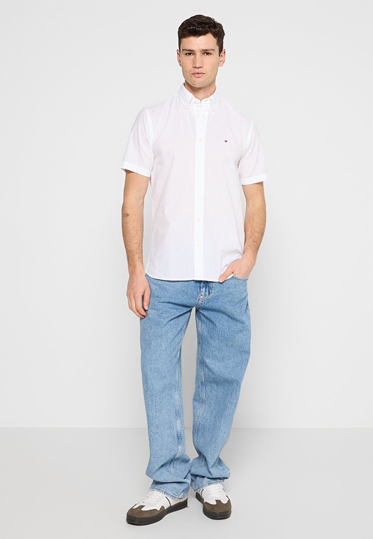Chemise blanche à rayures à manches courtes avec col boutonné, associée à un jean en denim bleu clair ample et des baskets blanches avec des accents noirs.
