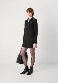Vestido polo preto de manga longa com golas, apresentando um detalhe de logotipo discreto, combinado com collants pretos transparentes e botins de salto agudo.
