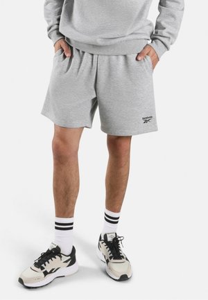 Person trägt hellgraue Reebok-Shorts mit den Händen in den Taschen, weiße Knöchelsocken mit schwarzen Streifen und beige-schwarze Sneakers.