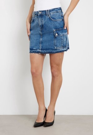 Mini-jupe en denim bleu avec poche à rabat latérale, ourlet effiloché, portée avec des talons noirs à bout pointu et un haut blanc.