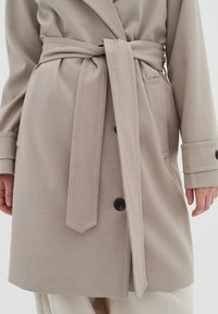 Beige kabát s ovázáním z měkké tkaniny, který má stahovací pásek, černé knoflíky a boční kapsy, má přizpůsobený střih a dlouhé rukávy.