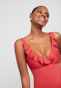 Maillot de bain rouge avec un décolleté en V profond et des accents froncés aux épaules. Texture lisse, design ajusté et sans motifs visibles.