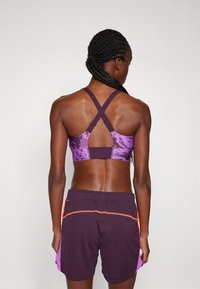 Violetti urheiluliivi, jossa on ristikkäinen selkäosa ja graafinen kuvio. Paritettu tummien shortsien kanssa, joihin on lisätty oranssit ja violetit sivupaneelit.