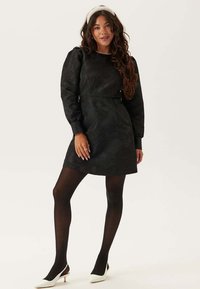 Abito nero con maniche a sbuffo, tessuto testurizzato e vita aderente. Indossato con collant neri e scarpe con tacco bianche. Accessorio semplice con fascia per capelli.