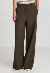 Pantalons larges en olive foncé, fabriqués à partir d'un tissu lisse. Présentent des poches avant, une taille ajustée et des chaussures pointues blanches.